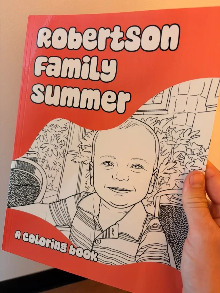 A hand holding the printed red Robertson Family Summer coloring book with a toddler portrait on the cover